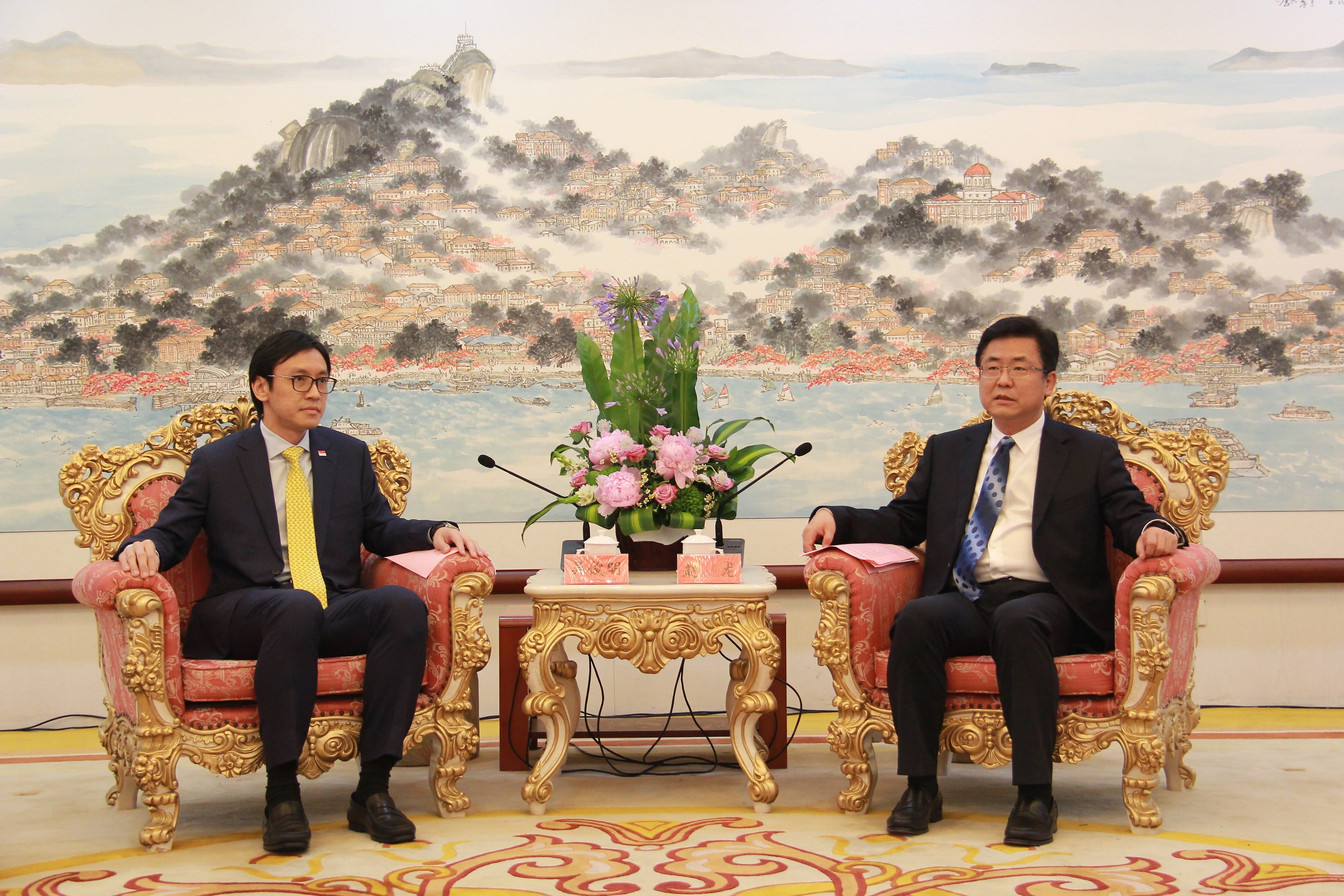 Two men in suits sit in ornate chairs against a landscape mural background.